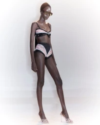 Black female model wearing a black and pale pink two-piece swimsuit, sunglasses, and strappy white heels against a white backdrop.