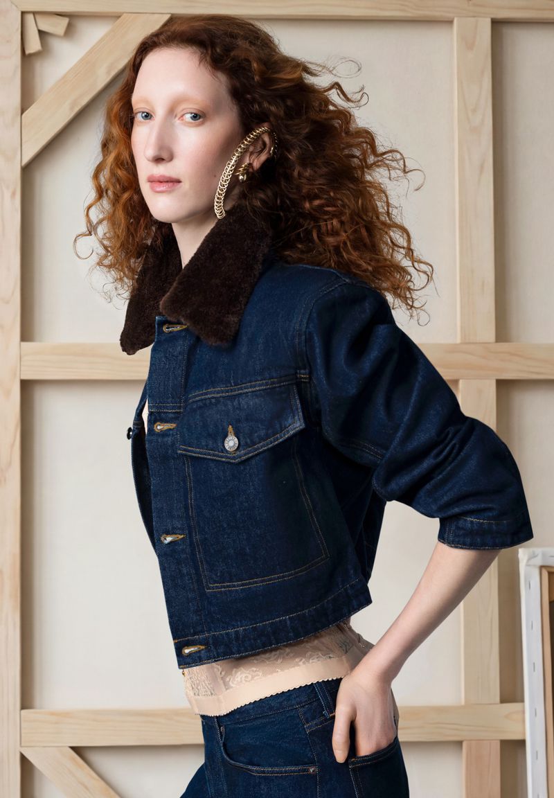 Model Lorna Foran posing in a dark blue denim jacket with a brown shearling collar against wooden canvas stretchers.