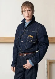 Model Thomas Westcott wearing a dark raw denim jacket embroidered with 'Dean' and matching denim trousers in a studio setting.