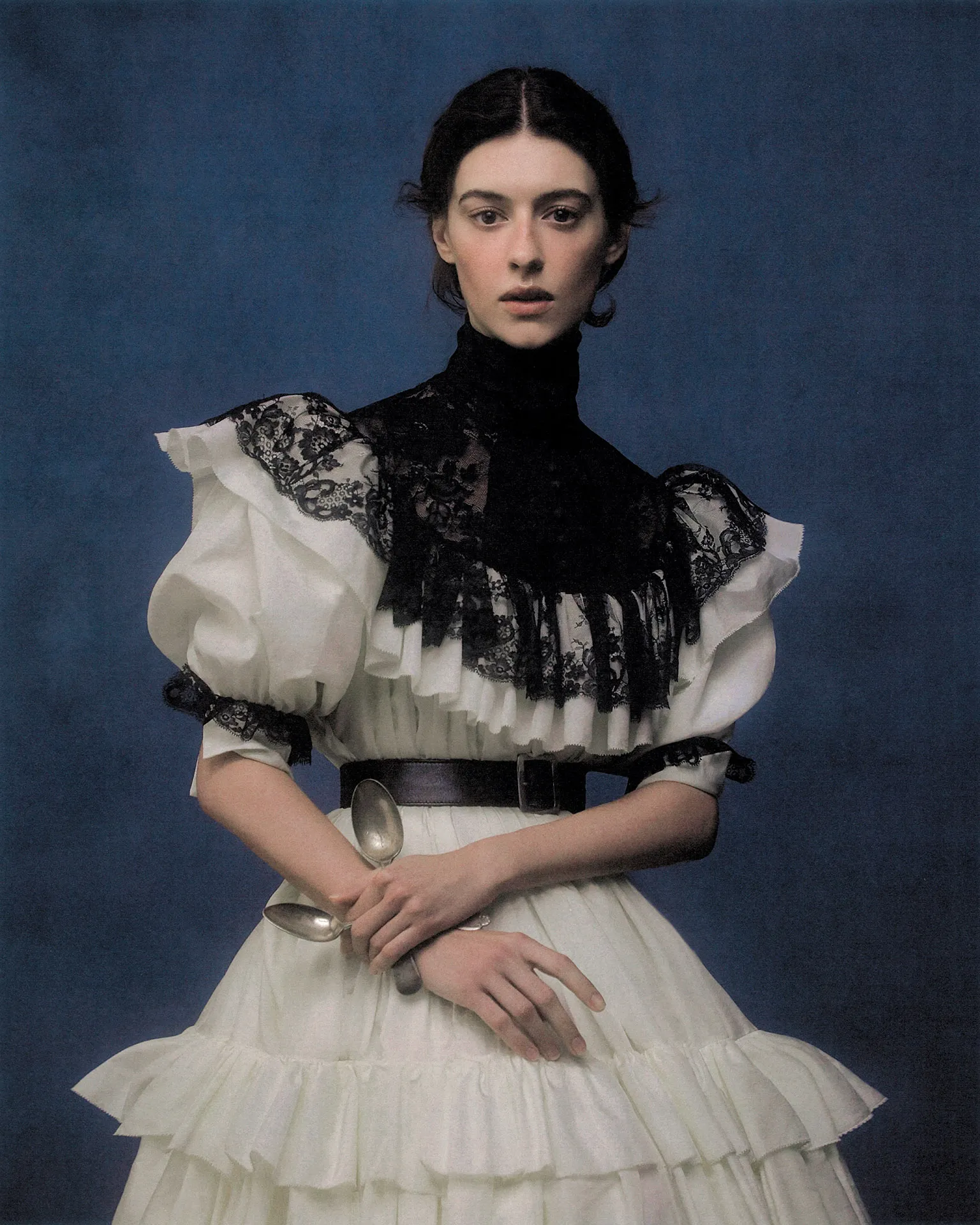 Daisy Edgar-Jones posing in a white ruffled dress with black lace trim and holding silver spoons against a dark blue textured backdrop.
