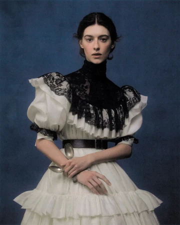 Daisy Edgar-Jones posing in a white ruffled dress with black lace trim and holding silver spoons against a dark blue textured backdrop.