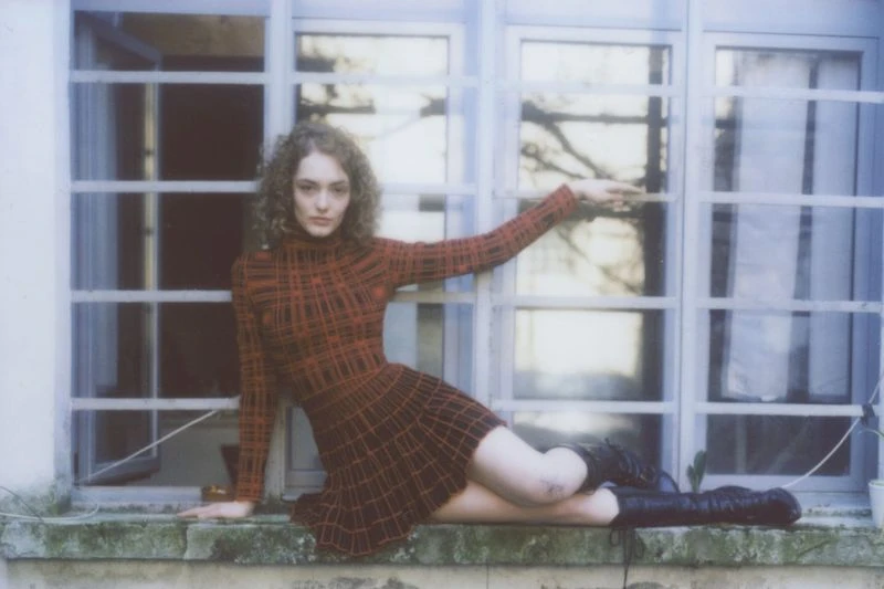 A woman with curly hair is sitting on a window sill in a red and black dress and black boots, with a window and tree visible in background.