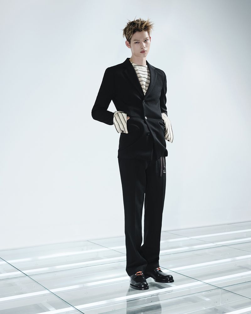 ENHYPEN member standing in a tailored black Prada suit and striped top on an illuminated floor against a white studio background.