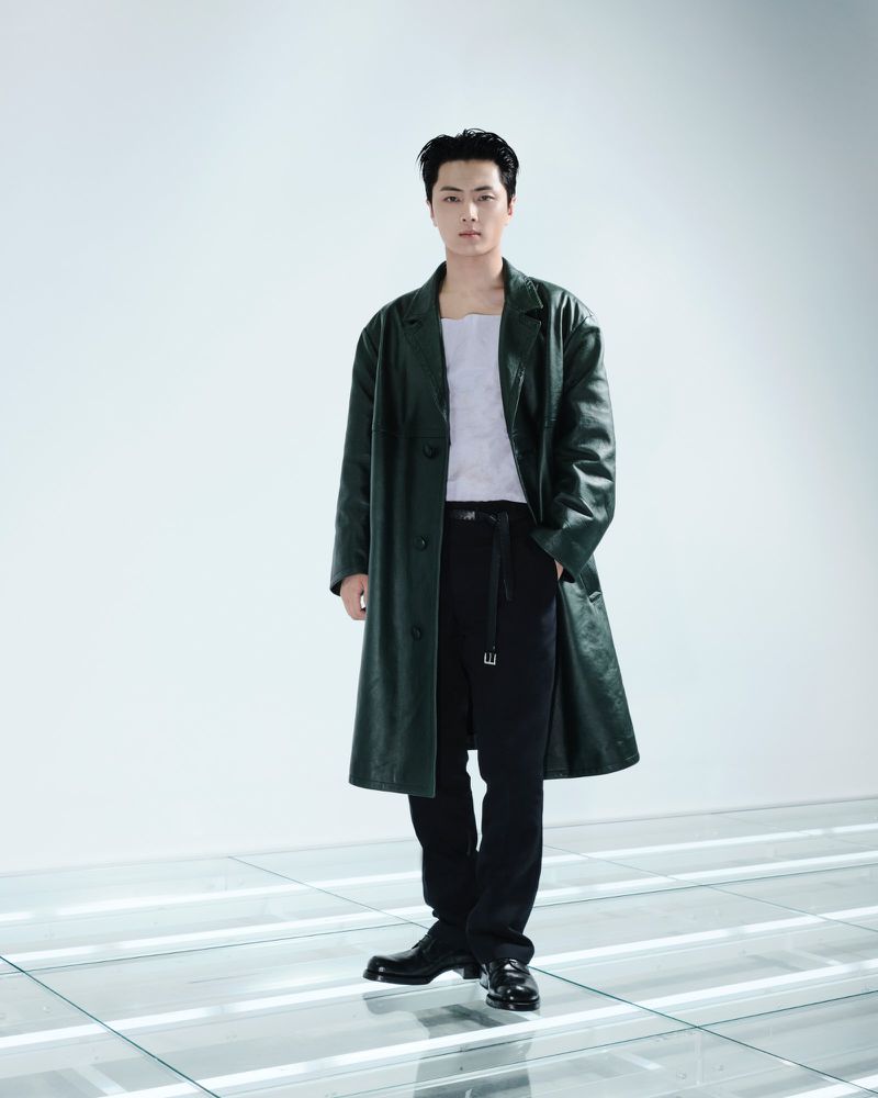 ENHYPEN's Jay standing on an illuminated glass floor wearing a dark green leather coat, white top, and black trousers for Prada.