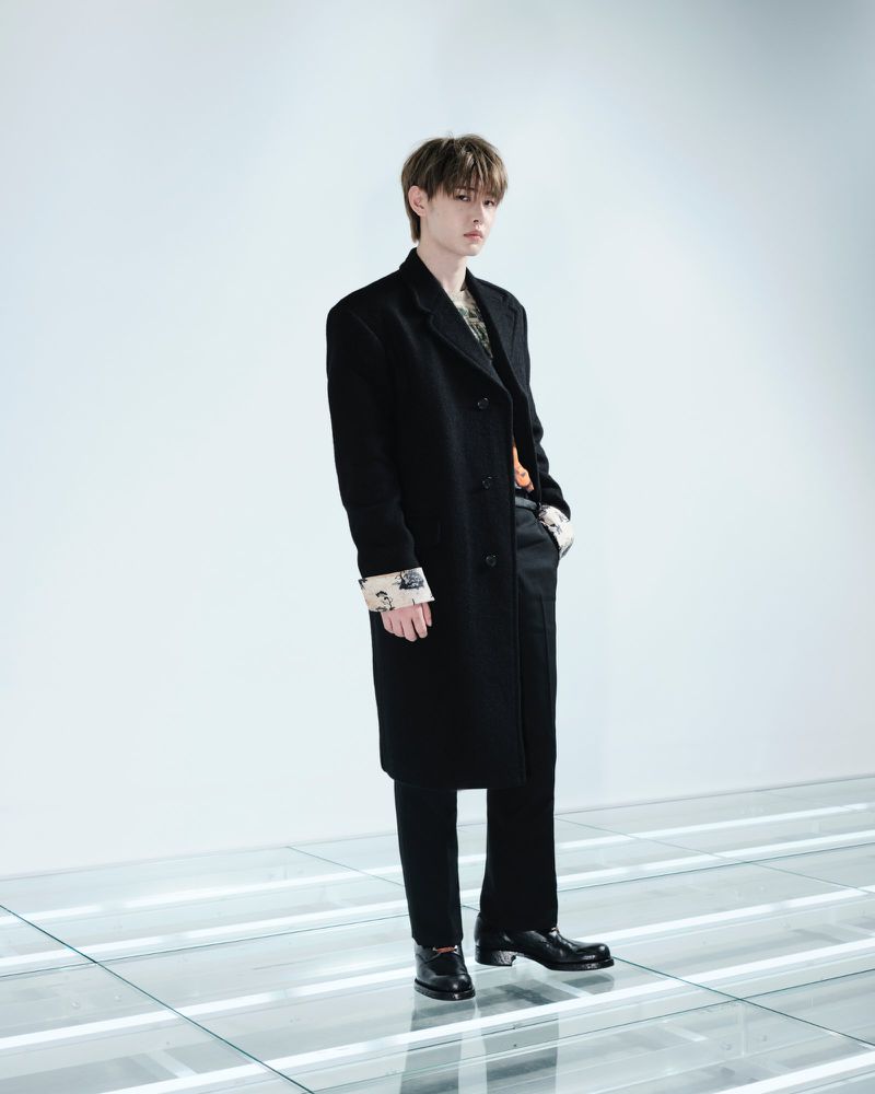 ENHYPEN member wearing a long black Prada overcoat and black dress shoes standing on an illuminated floor grid.