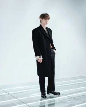 ENHYPEN member wearing a long black Prada overcoat and black dress shoes standing on an illuminated floor grid.