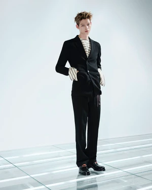 ENHYPEN member standing in a tailored black Prada suit and striped top on an illuminated floor against a white studio background.