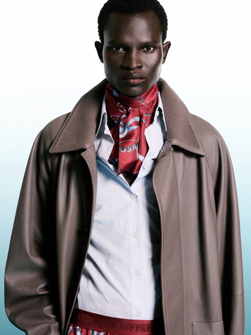 Model Chubath Kutien wearing a taupe leather coat, striped shirt, and red printed neck scarf against a gradient background.
