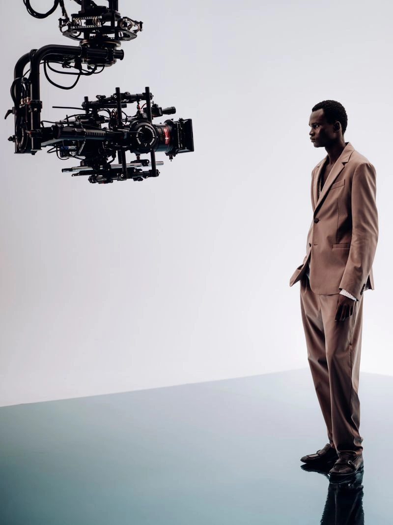 Model Chubath Kutien in a tan suit stands facing a suspended cinema camera on a studio set.