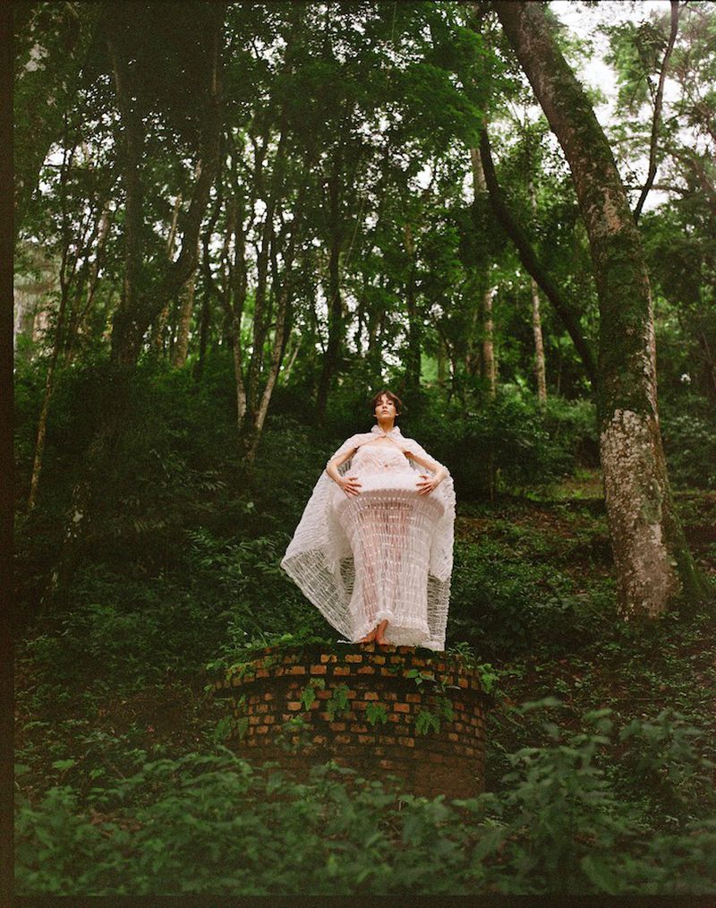 Woman in a white gown poses on a brick structure in a lush green forest setting, surrounded by trees and vegetation in natural light.