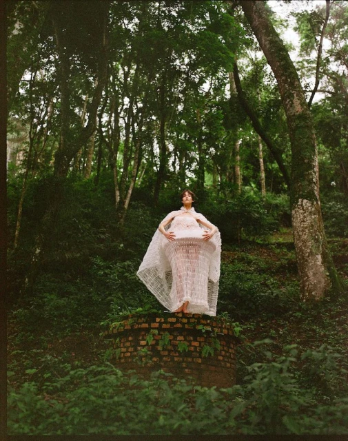 Woman in a white gown poses on a brick structure in a lush green forest setting, surrounded by trees and vegetation in natural light.