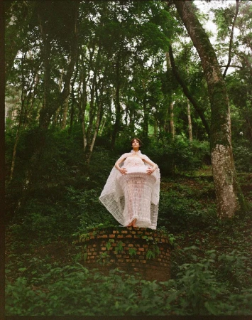 Woman in a white gown poses on a brick structure in a lush green forest setting, surrounded by trees and vegetation in natural light.