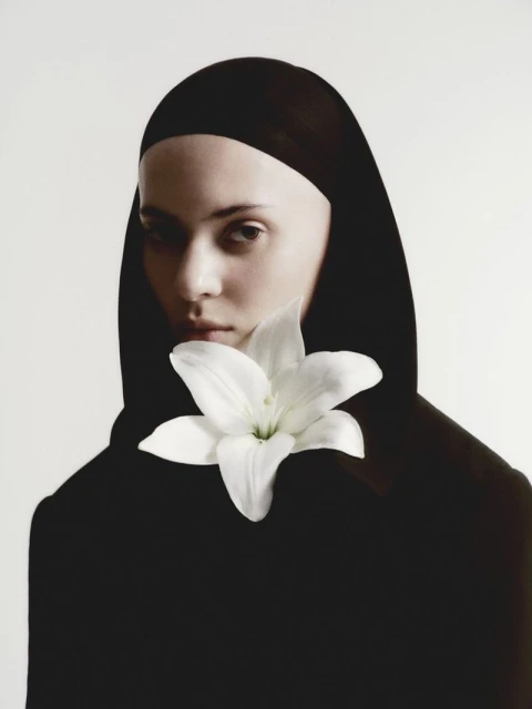 Portrait of a young woman wearing a black head covering and holding a white lily flower, set against a clean, bright white background.