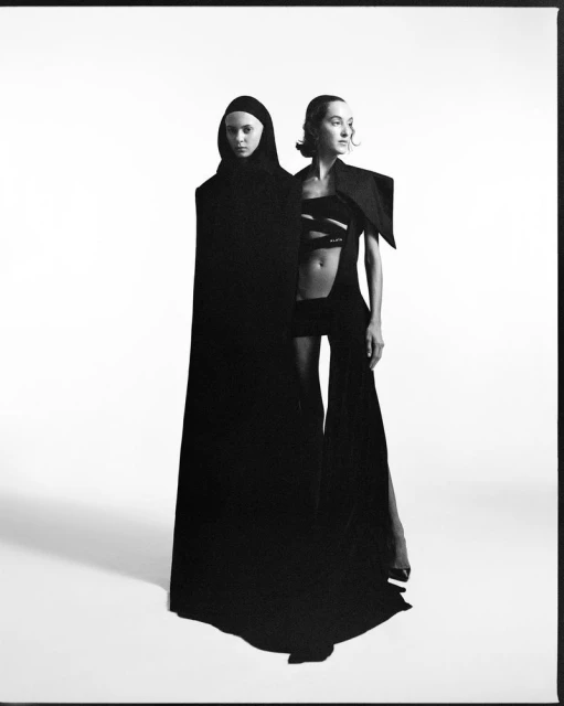 Two fashion models in black avant-garde hooded and cutout garments pose in a studio setting with stark lighting, creating a dramatic contrast.