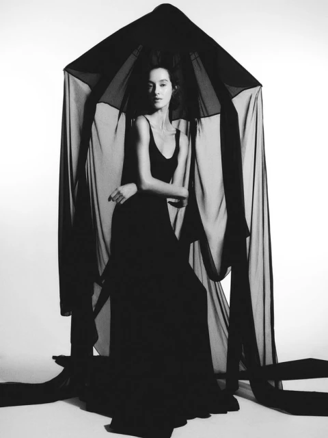 Woman in a long black dress and dark draping stands in a photo studio with bright white background, creating a contrast in colors.