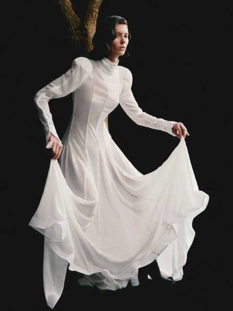 A slender woman in a white, flowing gown poses in a dark environment near a light-colored tree branch, captured with a neutral expression.