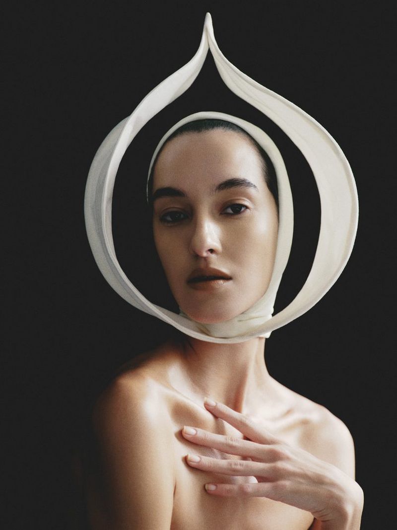 Close-up portrait of a bare-shouldered woman with hand on chest, wearing a white headpiece, against a dark background in a photo studio.