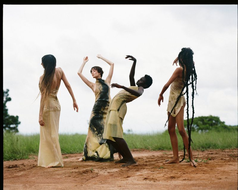Four women in pale dresses pose barefoot on a dirt path in a grassy field under a cloudy sky, showcasing elegant contemporary fashion.
