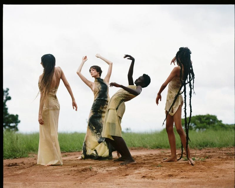 Four women in pale dresses pose barefoot on a dirt path in a grassy field under a cloudy sky, showcasing elegant contemporary fashion.