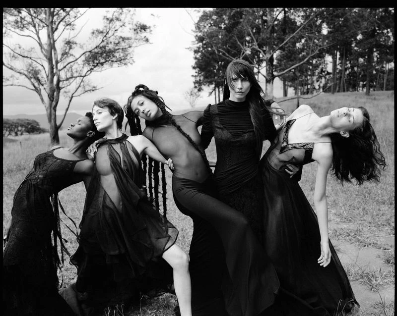 Black and white fashion shot featuring five women in flowing black dresses posing in an outdoor field with trees in the background.