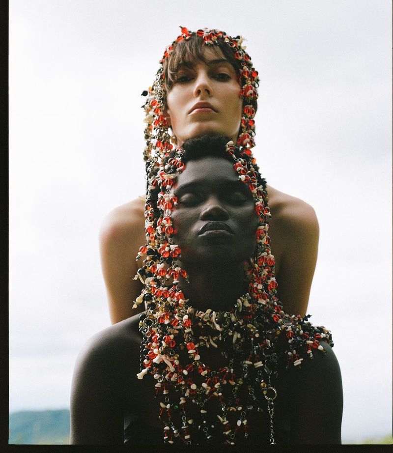 Two models, one Black and one White, pose in a studio wearing floral beaded headpieces and jewelry, with a blurred nature background.