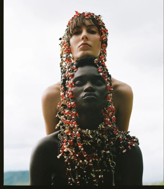 Two models, one Black and one White, pose in a studio wearing floral beaded headpieces and jewelry, with a blurred nature background.