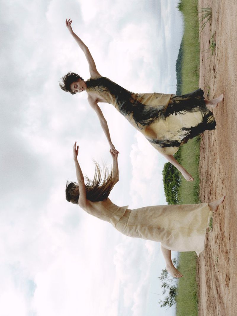 Two women in flowing dresses hold hands in a tilted view, with a sky background and a dirt road and green foliage in the distance.