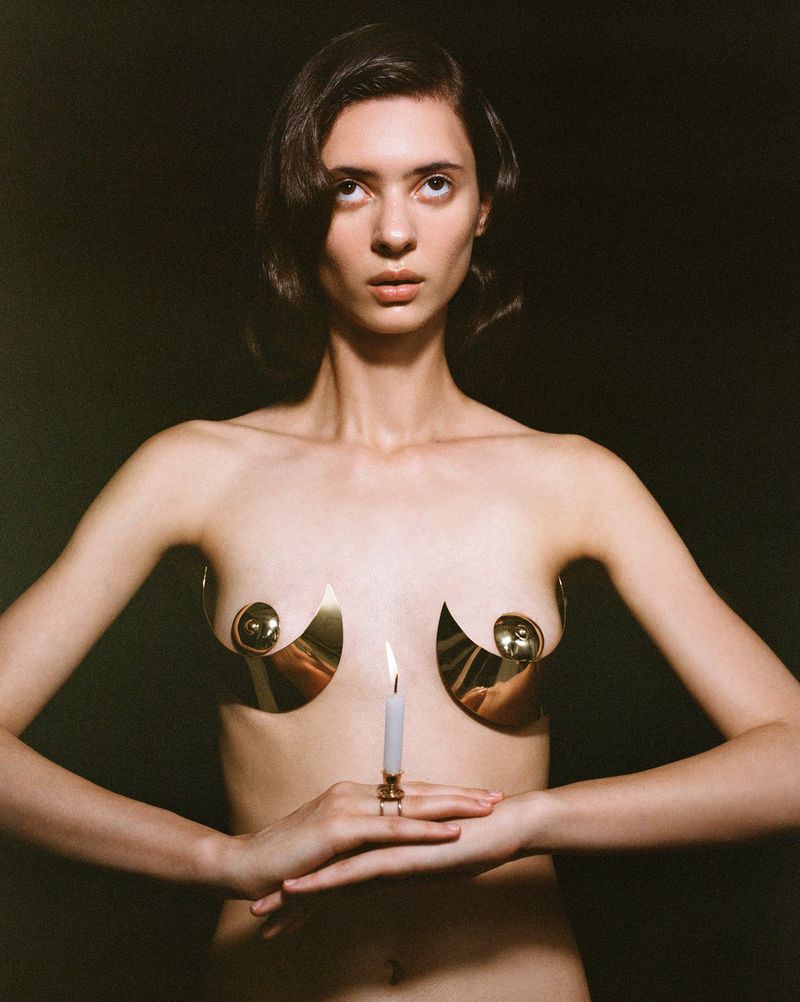 Woman poses in front of a dark background, wearing decorative gold-colored breastplates and holding a burning white candle.