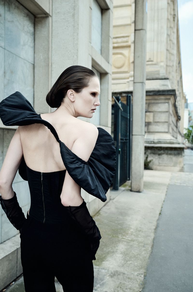 Model Alla Kostromichova poses in a black strapless sculptural top with draped sleeves on an urban sidewalk.