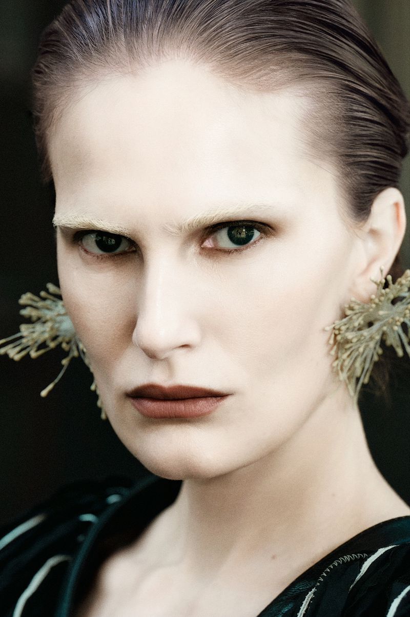 Close-up portrait of model Alla Kostromichova with bleached eyebrows, slicked-back hair, and sculptural earrings against a dark background.