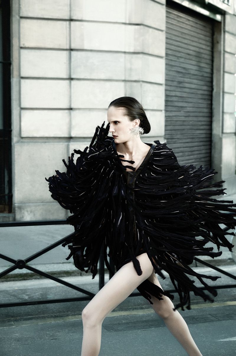Model Alla Kostromichova wearing a dynamic black fringe coat while striding past a stone building exterior on a city street.