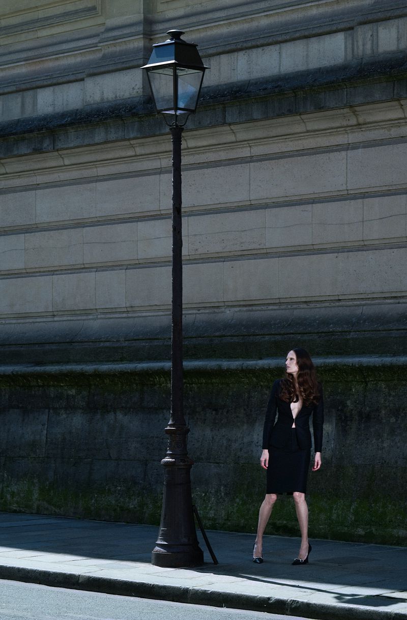 Alla Kostromichova in a black skirt suit and high heels posing next to a vintage street lamp along a stone building wall.