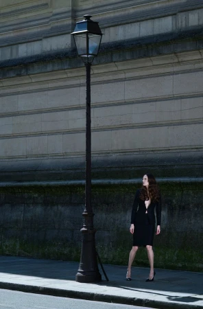 Alla Kostromichova in a black skirt suit and high heels posing next to a vintage street lamp along a stone building wall.