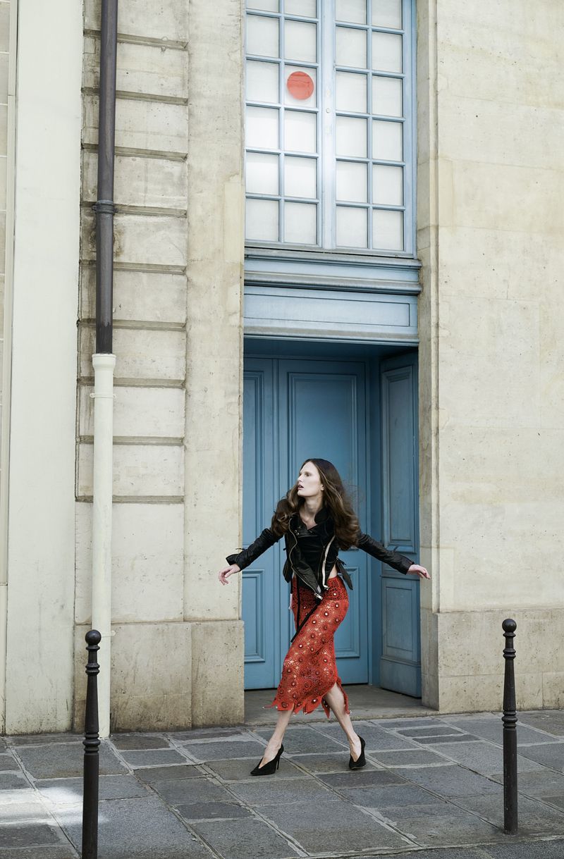 Alla Kostromichova poses dynamically in a black jacket and patterned rust skirt in front of tall blue doors on a stone sidewalk.
