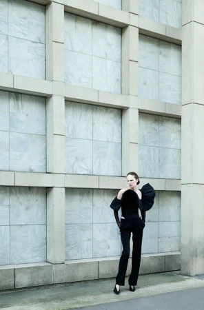 Model Alla Kostromichova posing outdoors in a sculptural black off-the-shoulder top and trousers against a grid wall.