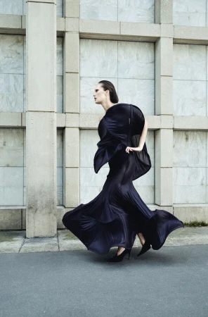 Alla Kostromichova walking in a sculptural black flowing gown and heels against an architectural grid facade.