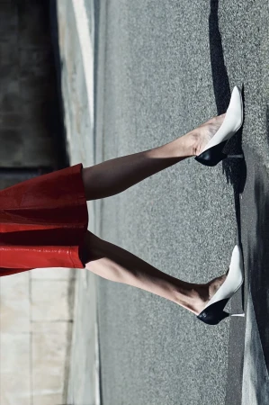 Model wearing a red skirt and two-tone black and white pointed-toe stiletto heels walking on an asphalt street in bright sunlight.