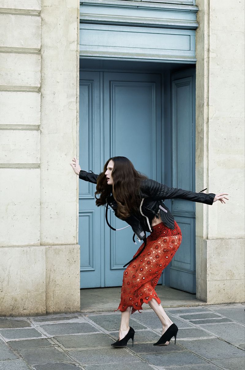Model Alla Kostromichova posing in front of a blue double door wearing a black jacket and red patterned skirt
