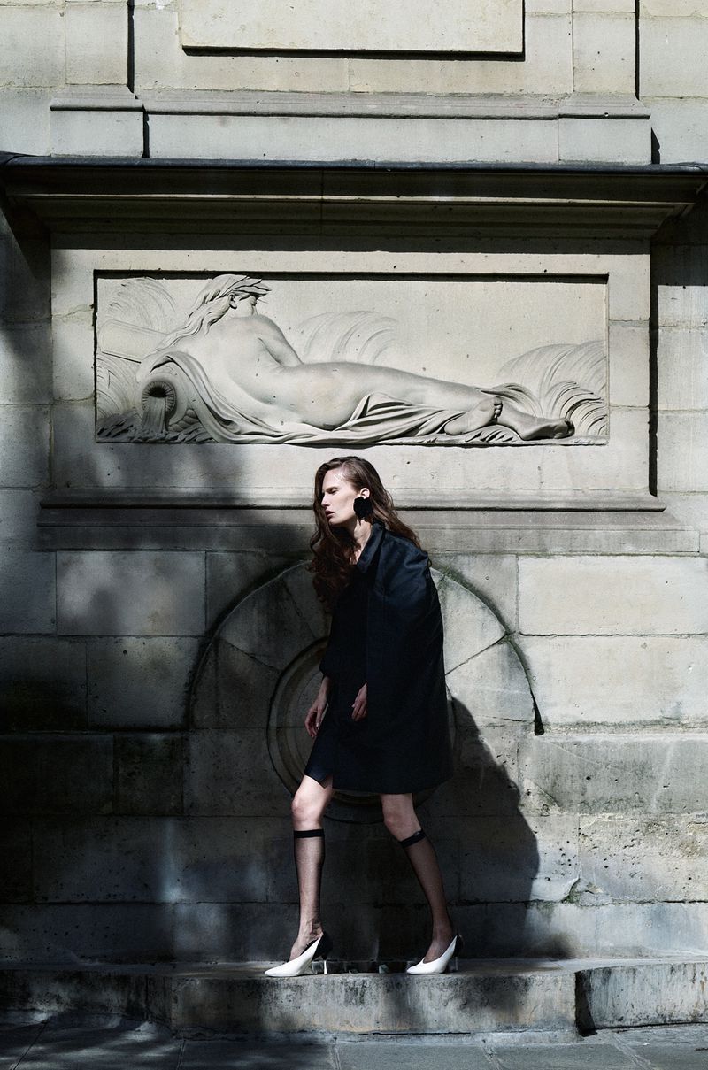Model Alla Kostromichova walking in a dark coat, sheer knee socks, and white heels in front of a stone wall with a classical relief sculpture.