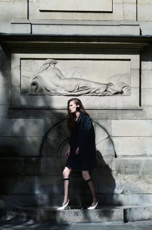 Model Alla Kostromichova walking in a dark coat, sheer knee socks, and white heels in front of a stone wall with a classical relief sculpture.