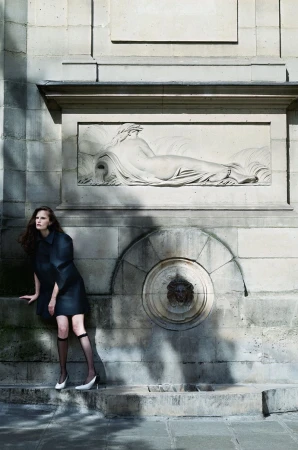 Alla Kostromichova posing in a black coat dress and knee-high socks beside a stone relief fountain in an editorial shoot.