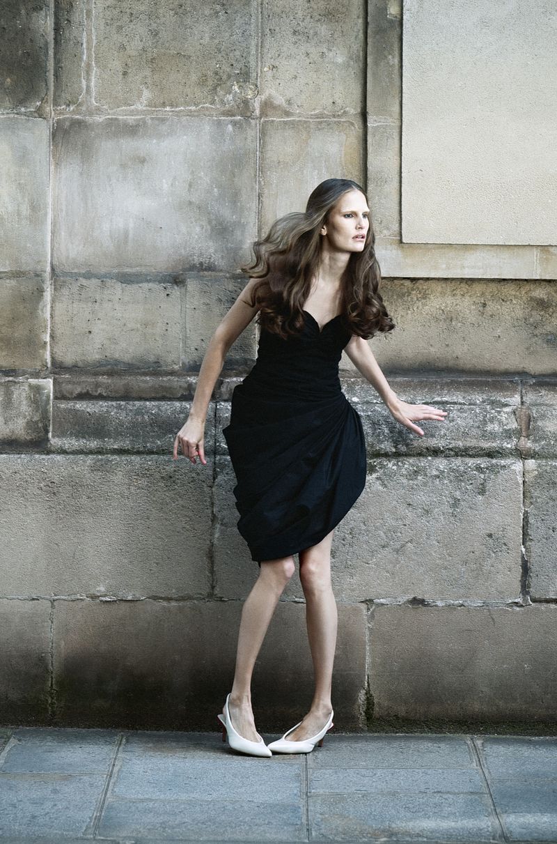 Model Alla Kostromichova wearing a strapless black draped dress and white slingback heels against a weathered stone wall.
