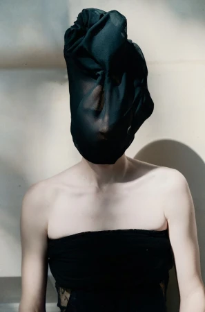 Model with head wrapped in sheer black fabric wearing a black strapless top against a neutral backdrop.
