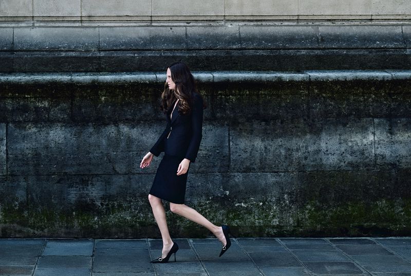 Model Alla Kostromichova walks along a stone wall wearing a tailored black skirt suit and pointed black high heels.