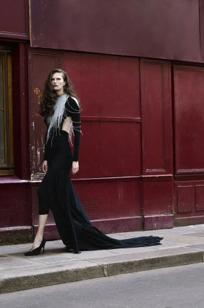 Model Alla Kostromichova walking along a sidewalk wearing a long black gown with silver fringe detail against a dark red building exterior.
