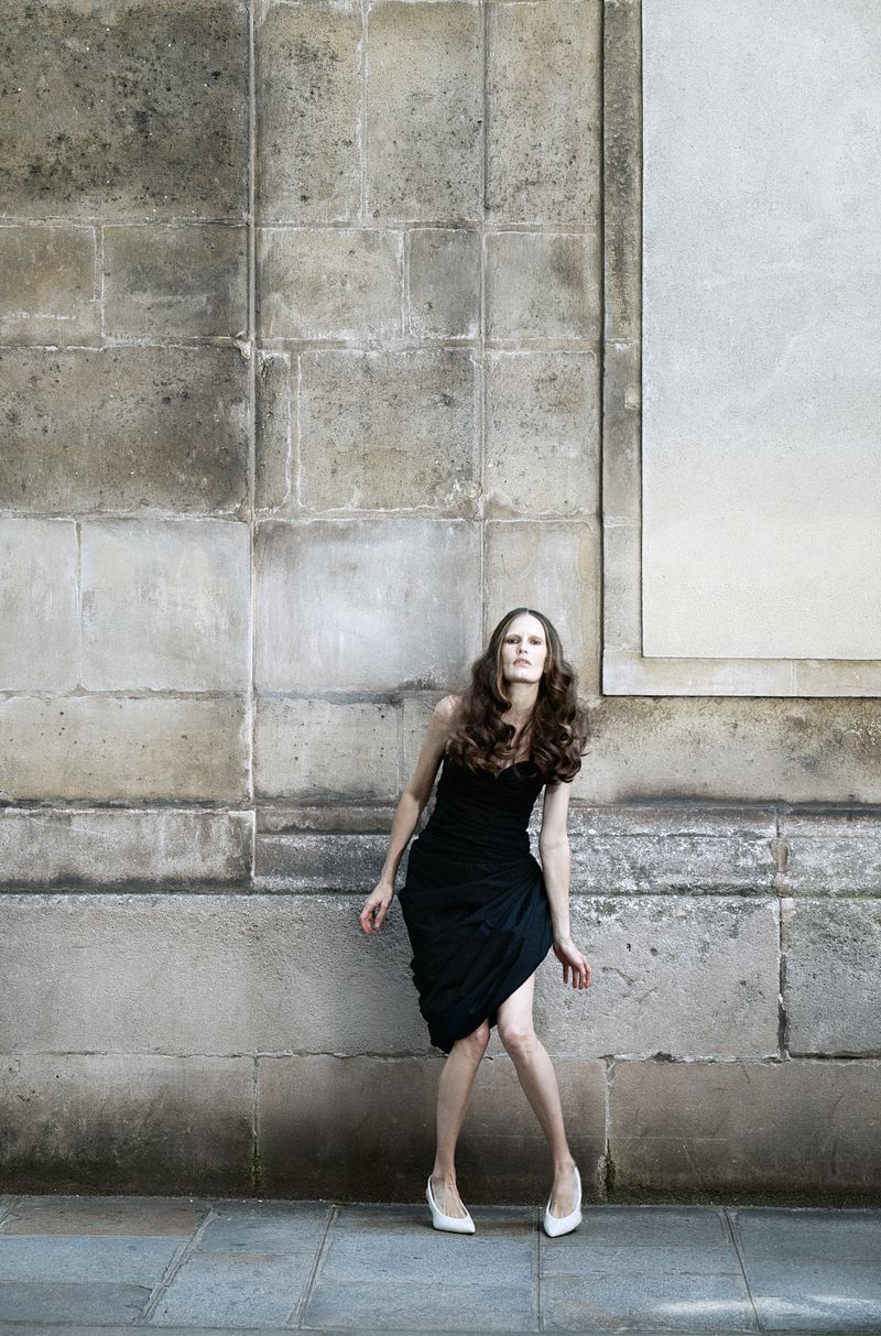 Model Alla Kostromichova wearing a black strapless draped dress and white pointed-toe pumps against a weathered stone wall.