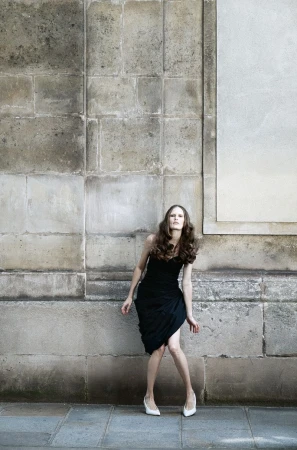 Model Alla Kostromichova wearing a black strapless draped dress and white pointed-toe pumps against a weathered stone wall.