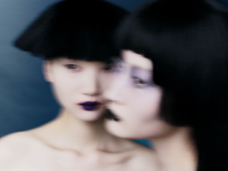 Two East Asian models with blunt black bob haircuts and dark purple lipstick in motion blur against a dark blue background.