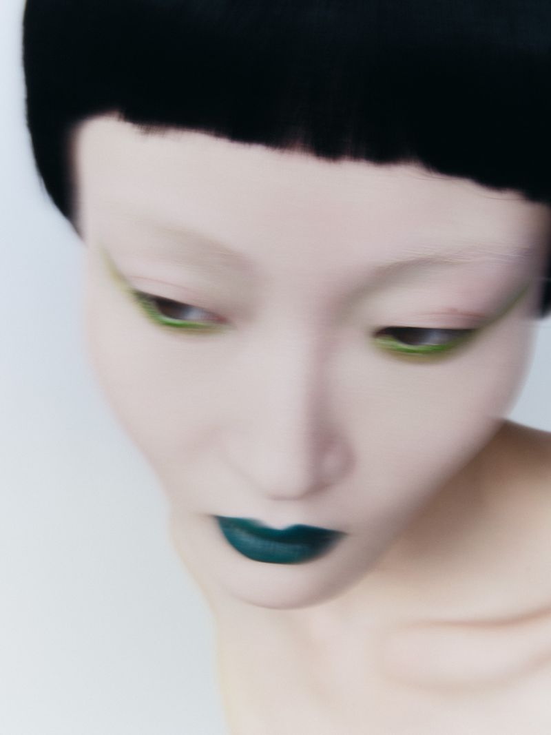 Close-up beauty portrait of an East Asian woman with blunt black hair, bold green eyeliner, and teal lipstick against a white backdrop.