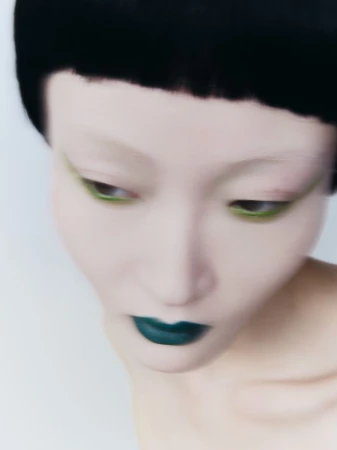 Close-up beauty portrait of an East Asian woman with blunt black hair, bold green eyeliner, and teal lipstick against a white backdrop.